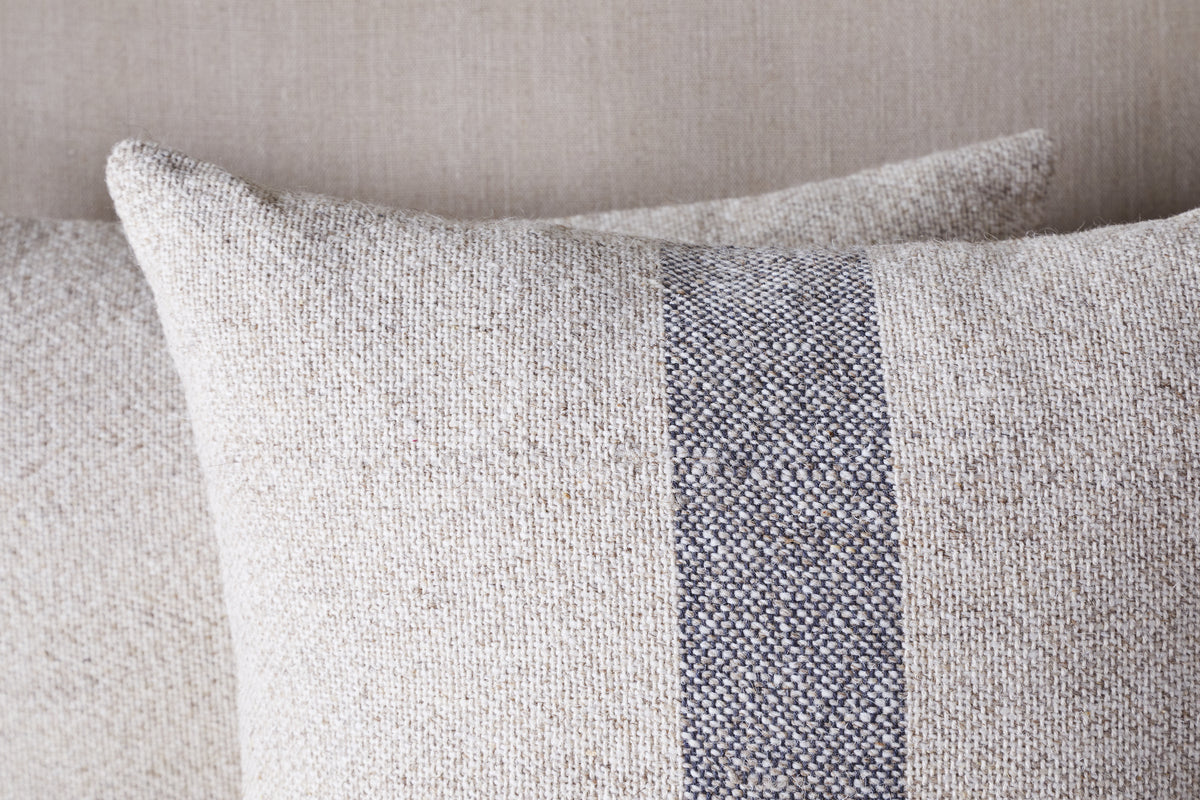 Two Kallu Wool & Cotton Cushion Covers by nkuku in natural tones, each with a central vertical blue-grey stripe, are arranged one behind the other against a matching fabric background.