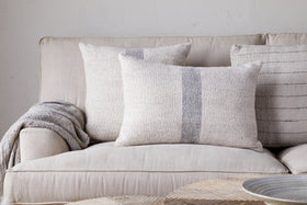 A beige sofa with nkuku’s Kallu Wool & Cotton Cushion Cover - Natural and three neutral throw pillows sits against a light wall, a gray knit blanket over the armrest. A round, light wood table appears in the foreground.
