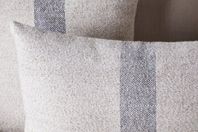 Two nkuku Kallu Wool & Cotton Cushion Covers in natural are shown overlapping, featuring a vertical blue-grey stripe pattern that highlights their woven texture and minimalist design.