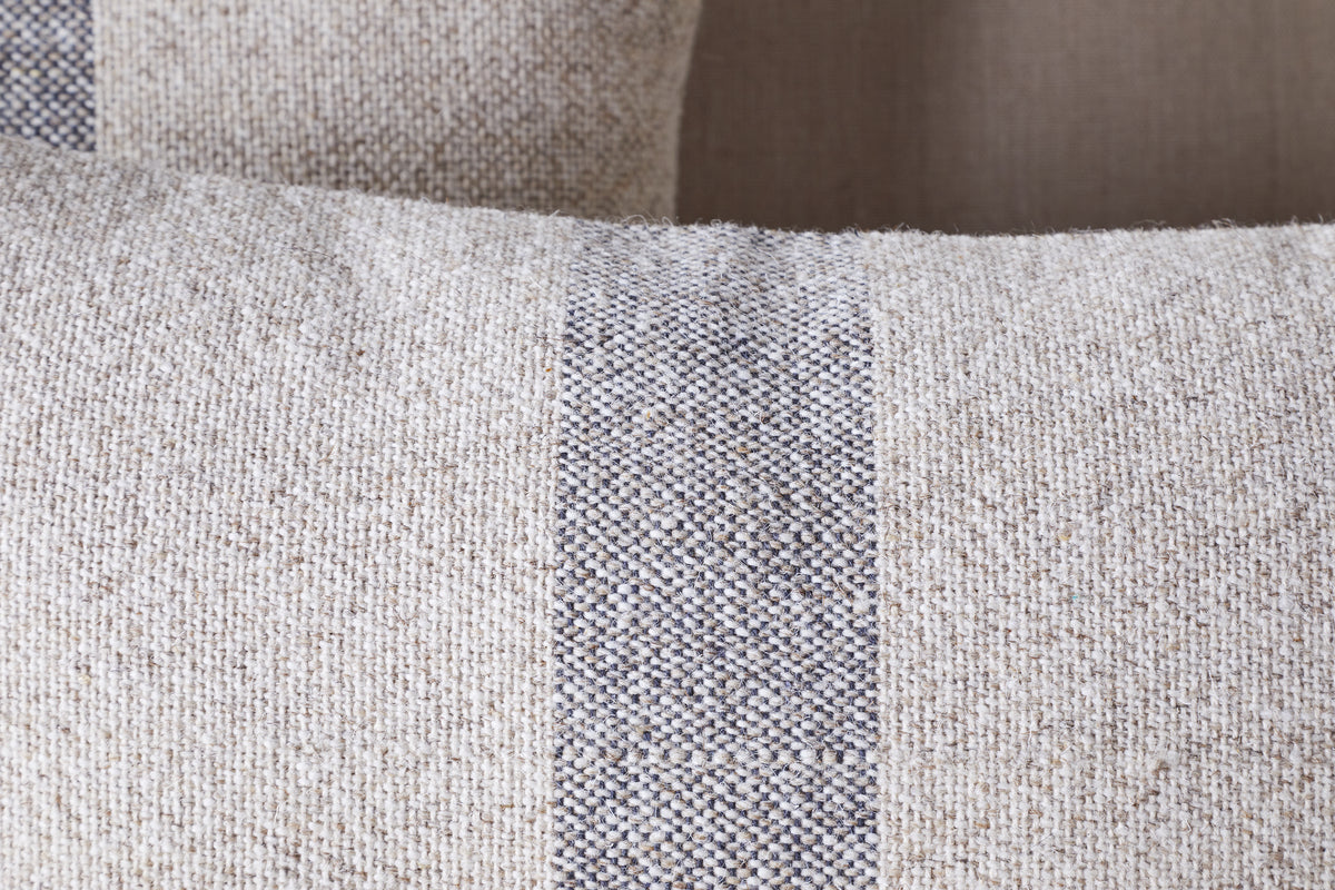 Close-up of the nkuku Kallu Wool & Cotton Cushion Cover - Natural, showing its textured beige fabric with a central vertical dark gray and white stripe. A similar cushion is softly blurred in the background.