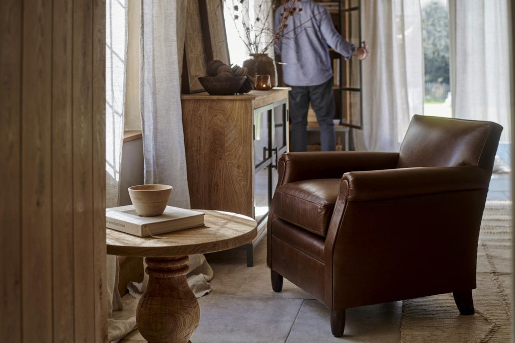 A cozy living room with the nkuku Knada Leather Armchair in chocolate brown, walnut-stained feet, a round wooden table with books and a bowl, and a wooden cabinet. A person stands by sheer curtains, looking outside through the glass door.