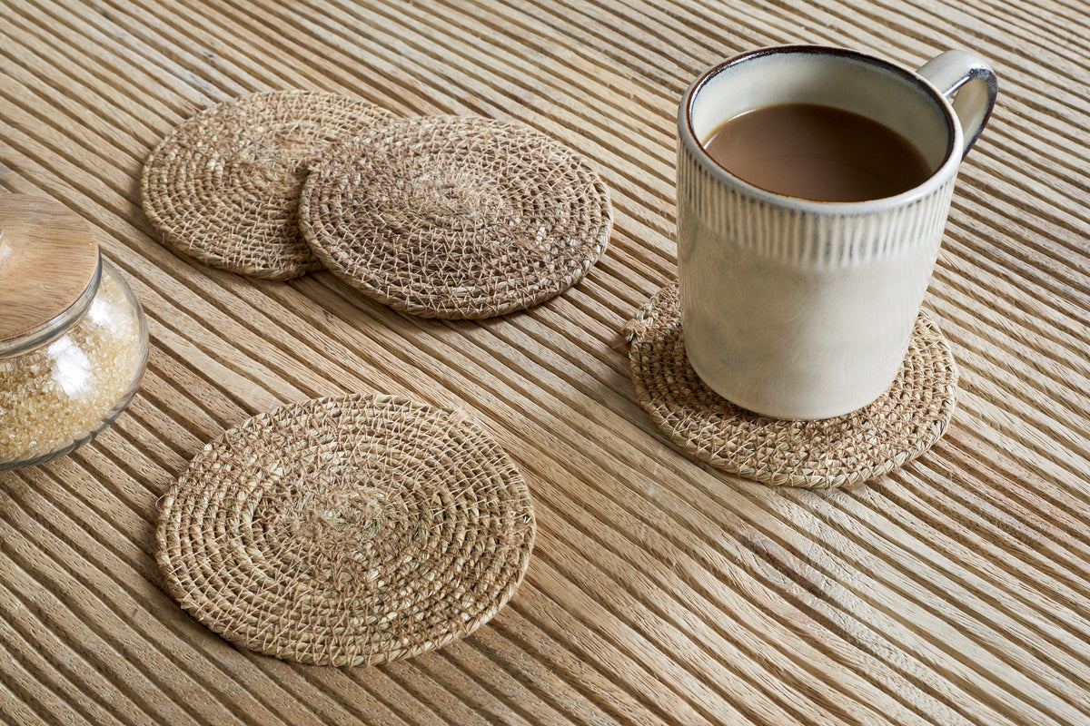A coffee-filled mug rests on an nkuku Keso Woven Coaster - Brown (Set of 4), with three more coasters and a glass jar of brown sugar with a wooden lid arranged on the textured wooden surface.