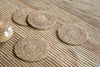 Four Keso Woven Coasters - Brown (Set of 4) by nkuku, handwoven from natural banana fibre, lie on a striped wooden surface. A jar with a wooden lid sits to the left, enhancing the earthy, minimalist tableware look.