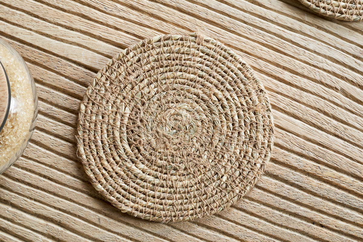 A Keso Woven Coaster - Brown from nkuku rests on a light wooden, striped surface, with part of a grain-filled glass container on the side, highlighting the minimalist style of this handwoven tableware set.