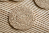 The Keso Woven Coasters - Brown (Set of 4) by nkuku, crafted from handwoven banana fibre, rest on a striped wooden surface. Another sleek tableware item is partly visible in the top right corner.