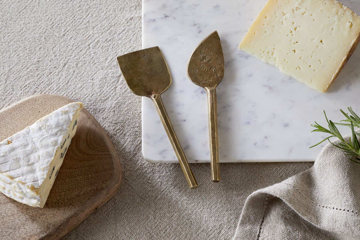 The nkuku Kasa Cheese Knives - Antique Brass (Set of 2) are displayed on a white marble board with hard cheese and brie on a wooden board, accompanied by a beige napkin and rosemary.