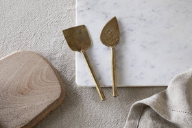 The nkuku Kasa Cheese Knives—antique brass, set of 2—rest on a white marble cheese board beside a wooden board and beige linen cloth, all arranged on a textured, light surface.