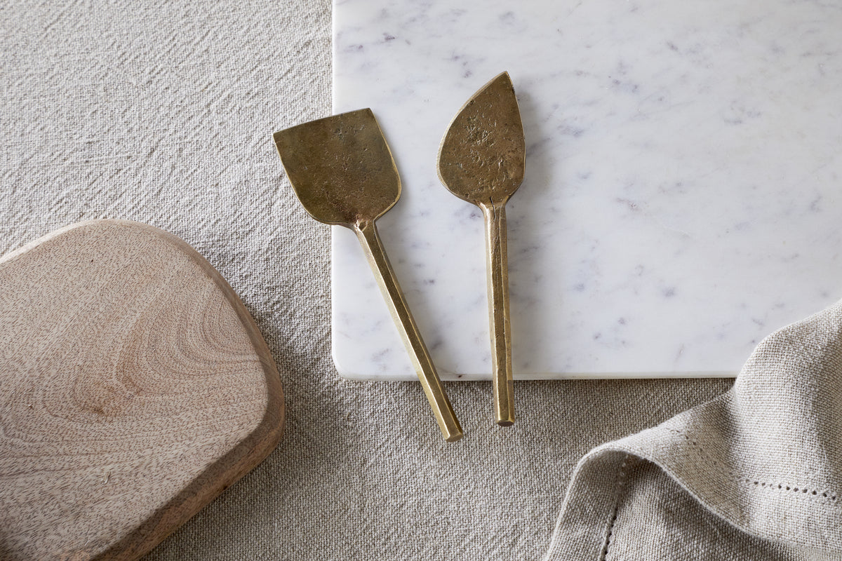 The nkuku Kasa Cheese Knives—antique brass, set of 2—rest on a white marble cheese board beside a wooden board and beige linen cloth, all arranged on a textured, light surface.