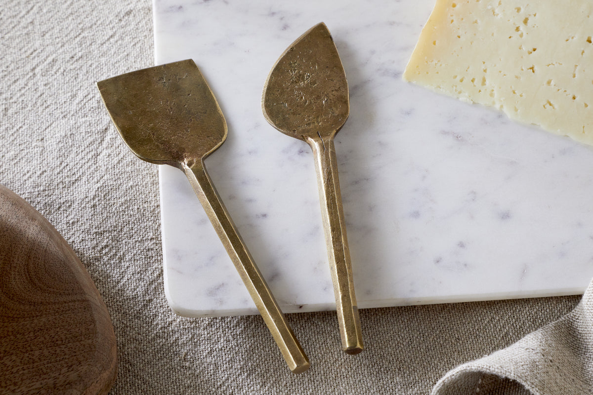 The Kasa Cheese Knives - Antique Brass (Set of 2) by nkuku rest on a white marble cheese board beside a wedge of cheese, with a beige textured cloth and part of a wooden bowl nearby.
