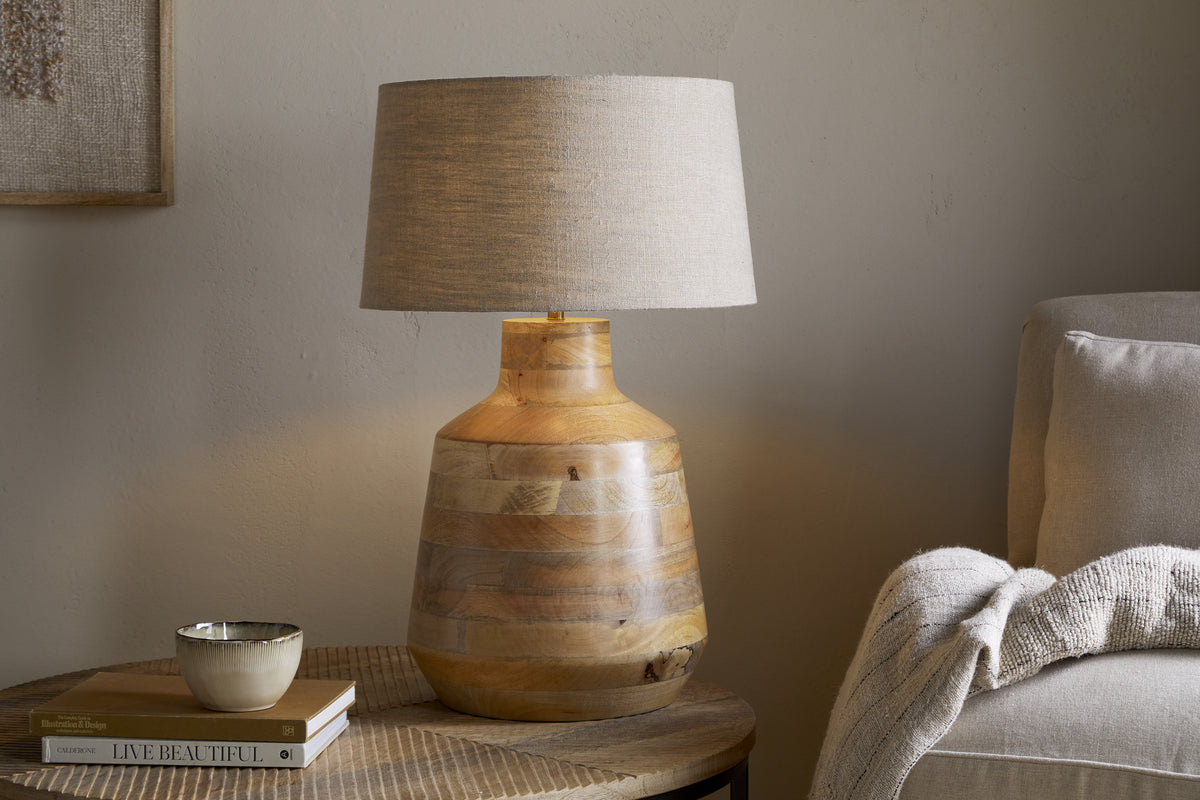 The Kayo Statement Table Lamp by nkuku, handcrafted from natural mango wood in India with a beige fabric shade, sits on a round table beside a folded blanket on a light sofa. A ceramic bowl and two books add to the cozy, neutral-toned setting.