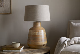 The nkuku Kayo Statement Table Lamp Mango Wood - Natural, handcrafted in India with a wooden base and beige shade, sits on a round side table beside books and décor—perfect statement lighting for cozy corners.