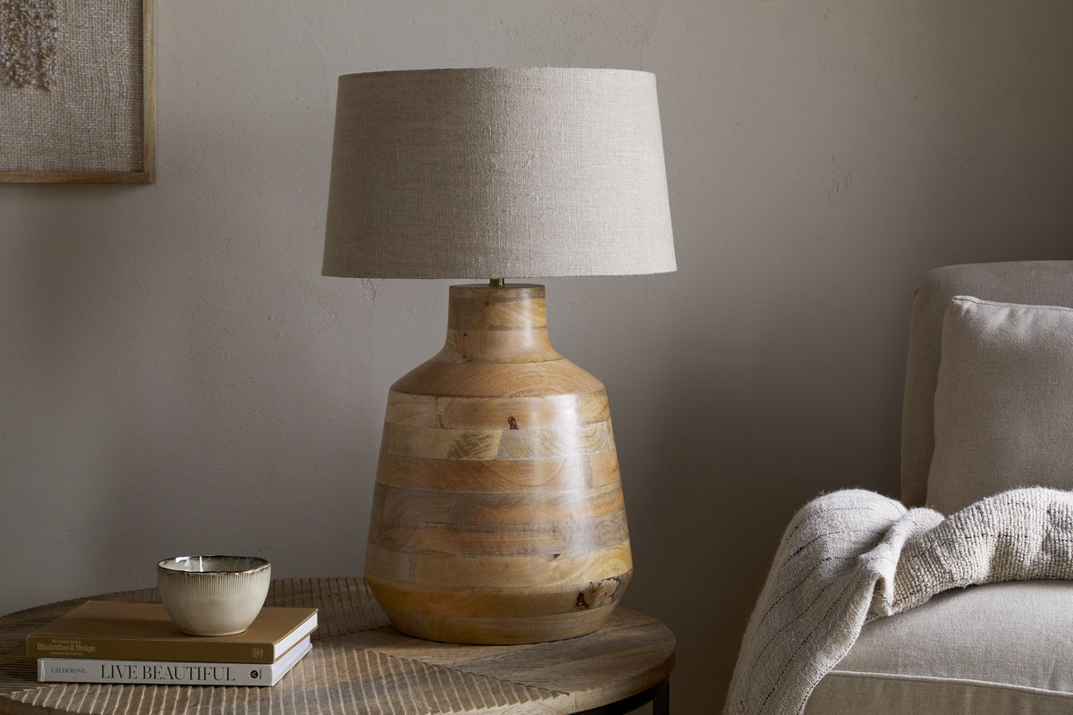 The nkuku Kayo Statement Table Lamp Mango Wood - Natural, handcrafted in India with a wooden base and beige shade, sits on a round side table beside books and décor—perfect statement lighting for cozy corners.