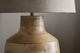 The nkuku Kayo Statement Table Lamp features a large beige linen shade and a natural mango wood base with a light grain, making it a standout piece against any plain, light-colored wall.