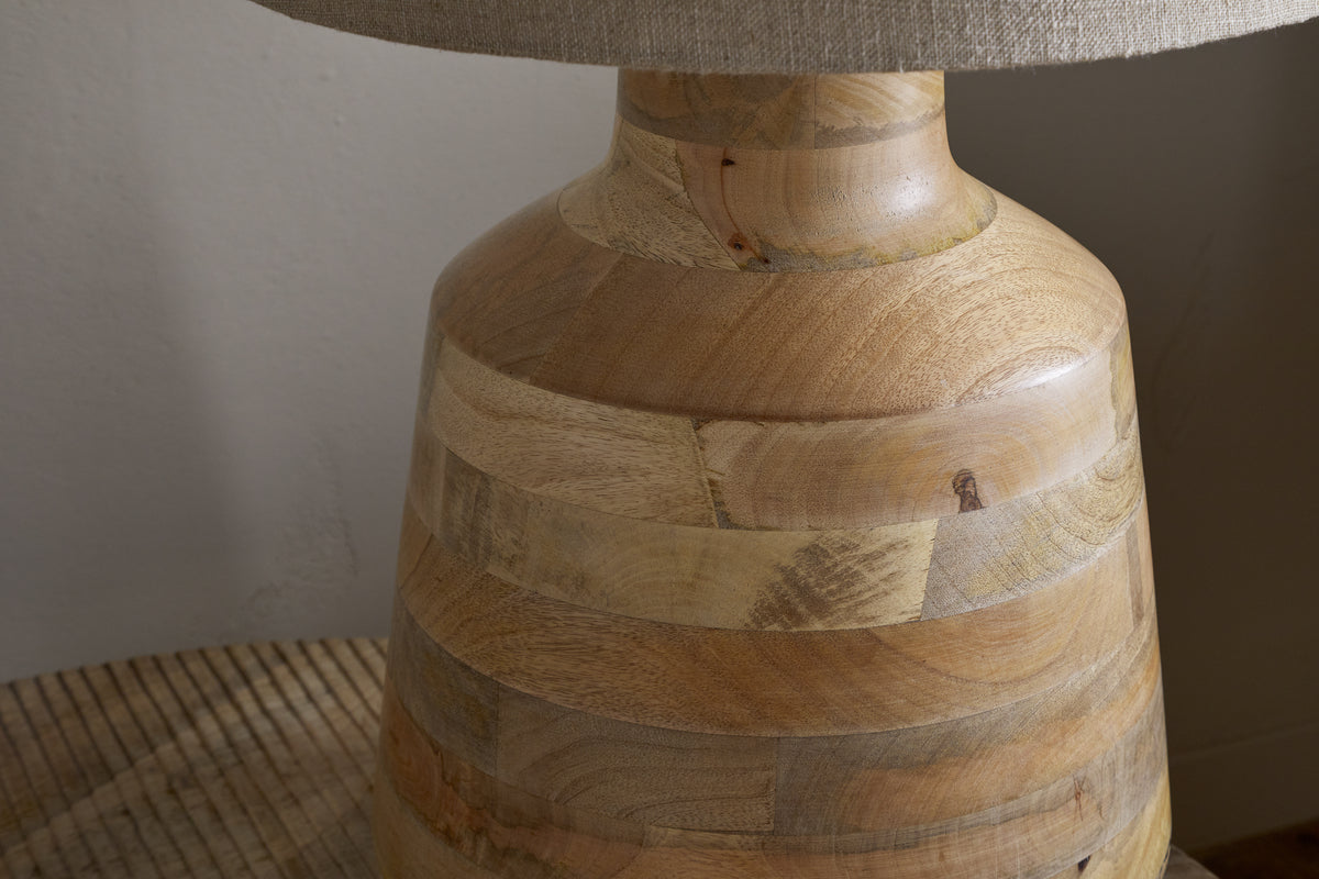 Close-up of the nkuku Kayo Statement Table Lamp in natural mango wood, handcrafted in India with a smooth finish and visible grain. The base rests on a wooden surface, while a beige fabric lampshade is partially visible above.
