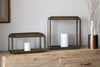Two nkuku Kiwanda Wide Lanterns in Antique Brass, each holding a white pillar candle, sit on a rustic wooden console table by a light wall. A tall vase with dried grasses completes this elegant candlelight display.