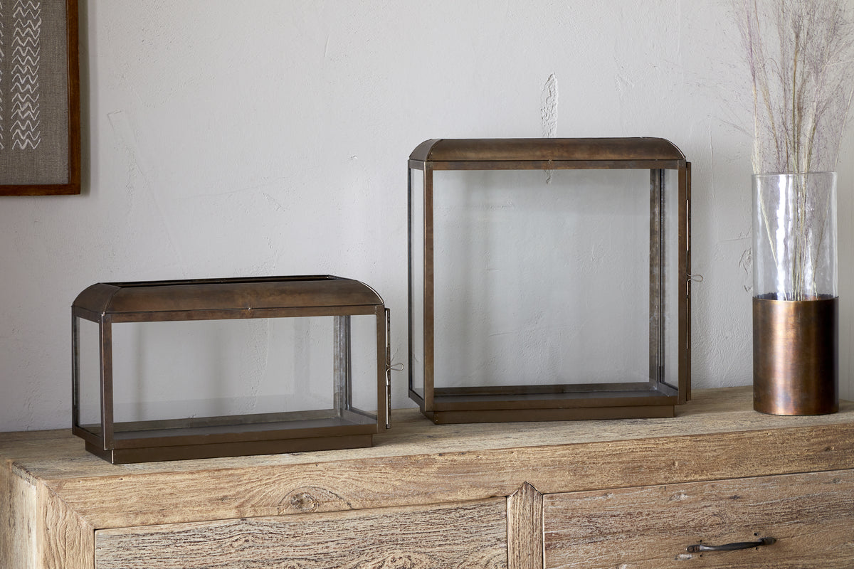 Two empty Kiwanda Wide Lanterns by nkuku, with antique brass frames, sit on a wooden sideboard. One rectangular and one square, they pair with a glass vase of dried grasses for modern candlelight décor.