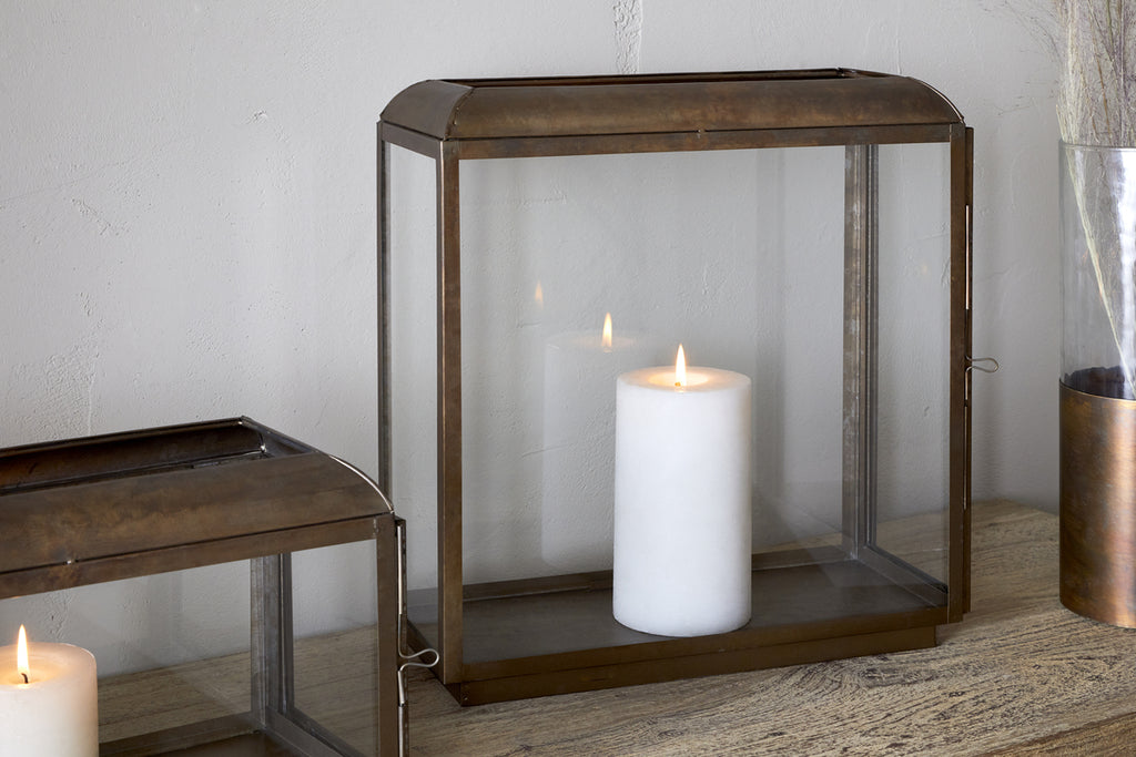 A lit white pillar candle glows inside the nkuku Kiwanda Tall Lantern in Antique Brass on a wooden surface, with another lantern and a vase of dried flowers nearby for an elegant candlelit display.