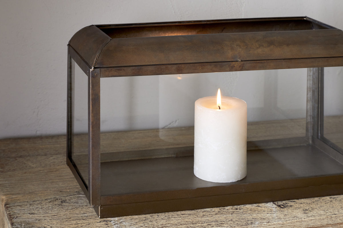 The Kiwanda Tall Lantern - Antique Brass by nkuku features a white pillar candle burning inside its rectangular metal and glass frame, adding timeless charm to any wooden surface and creating elegant candlelight against light-colored walls.