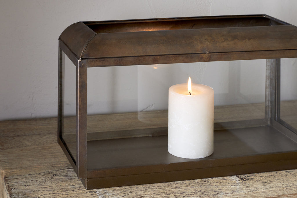 A lit white pillar candle glows inside the nkuku Kiwanda Wide Lantern in Antique Brass, placed on a wooden surface for elegant candlelight décor.