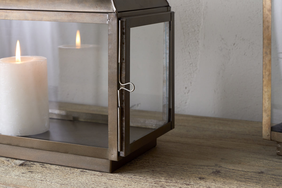 A white pillar candle glows inside the nkuku Kiwanda Tall Lantern in Antique Brass, adding candlelit decor to a wooden surface against a light-colored wall.