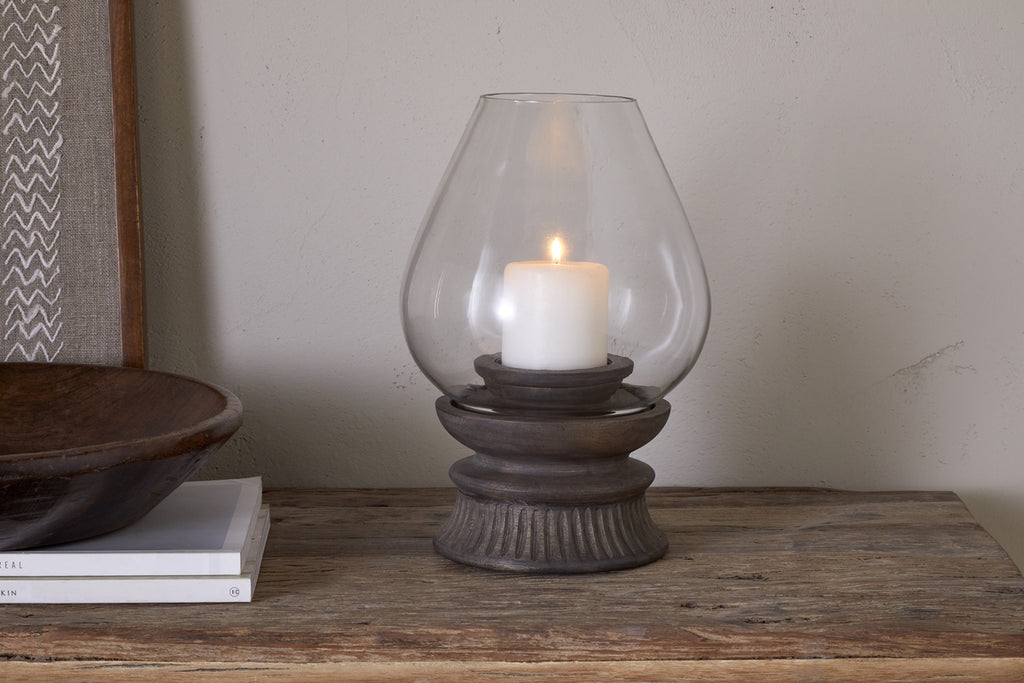 A lit white pillar candle sits inside the nkuku Kawani Mango Wood Lantern in Dark Ash, placed on a wooden table next to stacked books and a shallow wooden bowl.