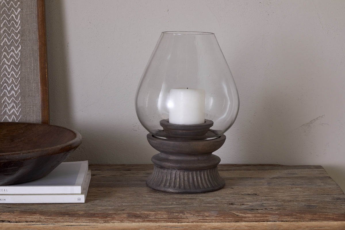 A white rustic pillar candle is placed inside the nkuku Kawani Mango Wood Lantern - Dark Ash on a wooden table, accompanied by stacked books and a wooden bowl.