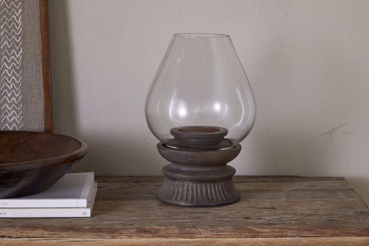The nkuku Kawani Mango Wood Lantern in Dark Ash rests on a rustic table beside a shallow wooden bowl and white books, perfect for displaying pillar candles in style.