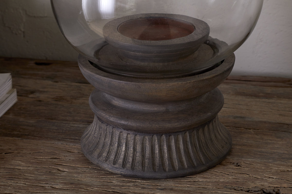 A close-up of the nkuku Kawani Mango Wood Lantern in Dark Ash, featuring rustic carved mango wood and a glass dome, sits on a weathered wooden surface beside a small stack of white paper.