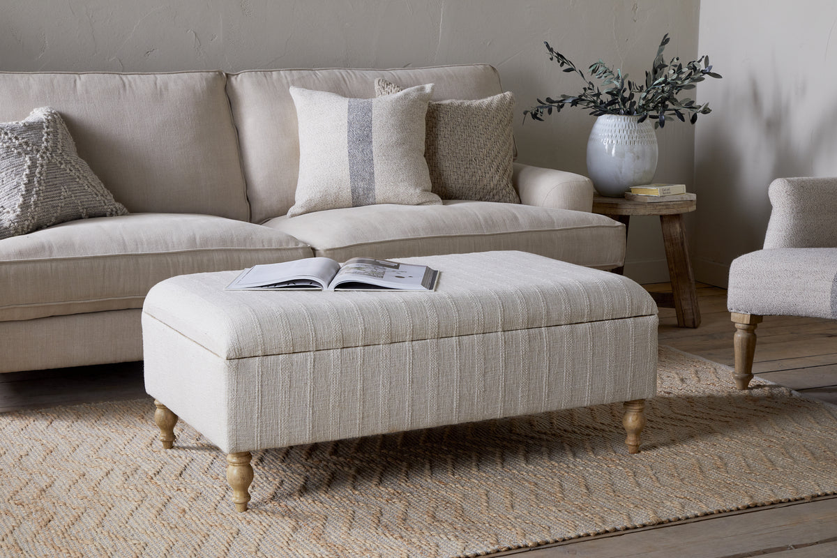 The Kiana Upholstered Storage Ottoman by nkuku in natural sits on a woven rug before a light sofa with neutral pillows. A round side table with a white vase and greenery completes this inviting living space.