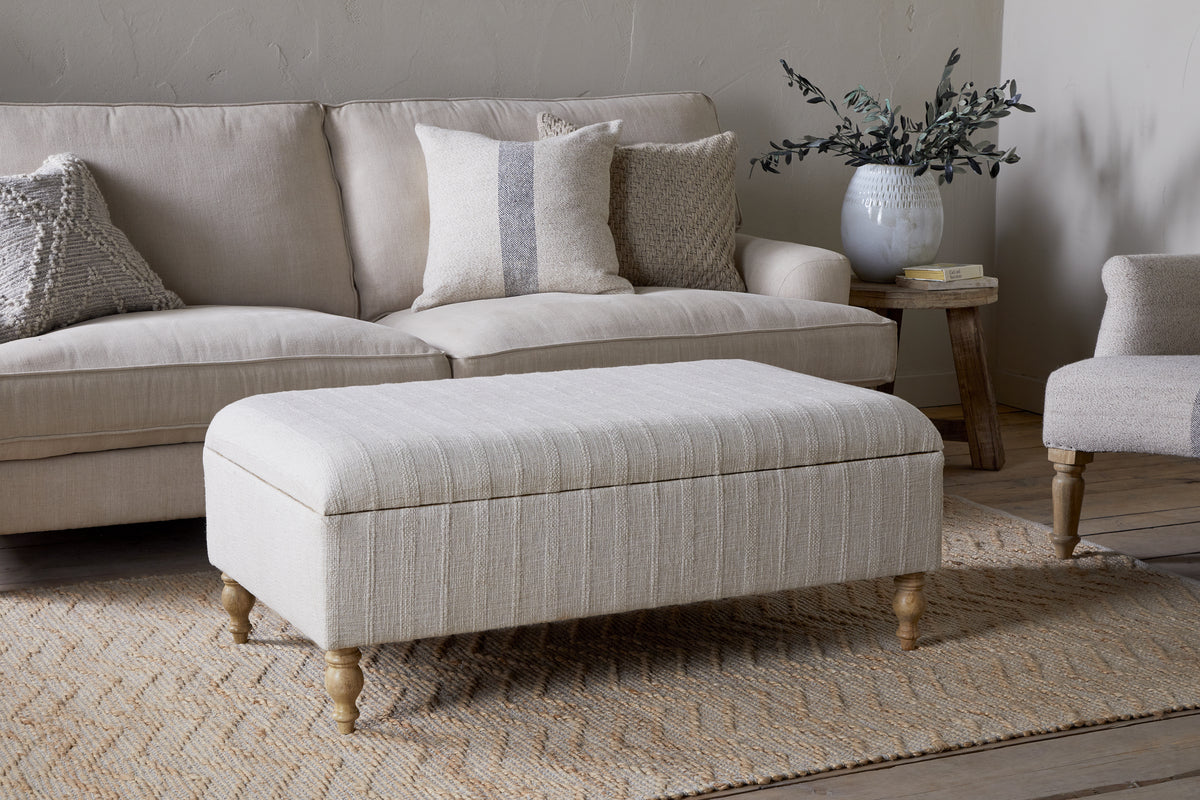 The Kiana Upholstered Storage Ottoman by nkuku in natural sits on a woven rug before a light sofa with pillows. Nearby, a side table holds a vase of greenery, completing the neutral-toned living space.