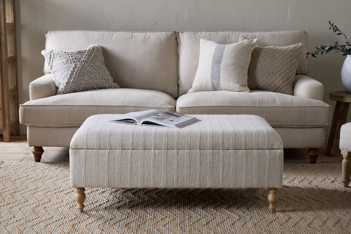 A beige sofa with four textured throw pillows sits behind the Kiana Upholstered Storage Ottoman by nkuku in Natural, which holds an open book on top, all arranged on a woven rug in a cozy, neutral-toned living space.