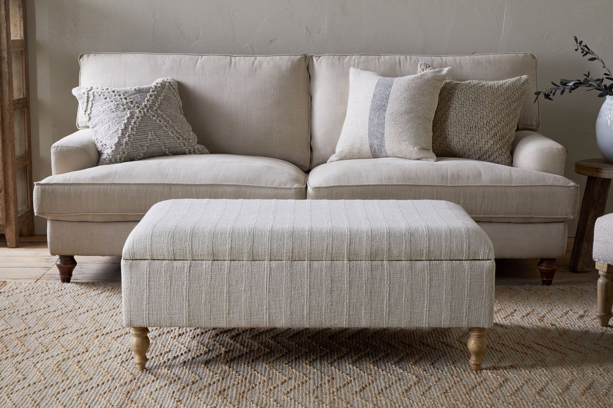 A nkuku Kiana Upholstered Storage Ottoman in natural sits before a beige sofa with three patterned throw pillows on a textured rug, creating a cozy, neutral-toned living space.