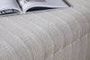 A close-up of the nkuku Kiana Upholstered Storage Ottoman in Natural, featuring a cream-striped, textured fabric and cushioned top, with an open magazine resting on it—an inviting accent for any living space.