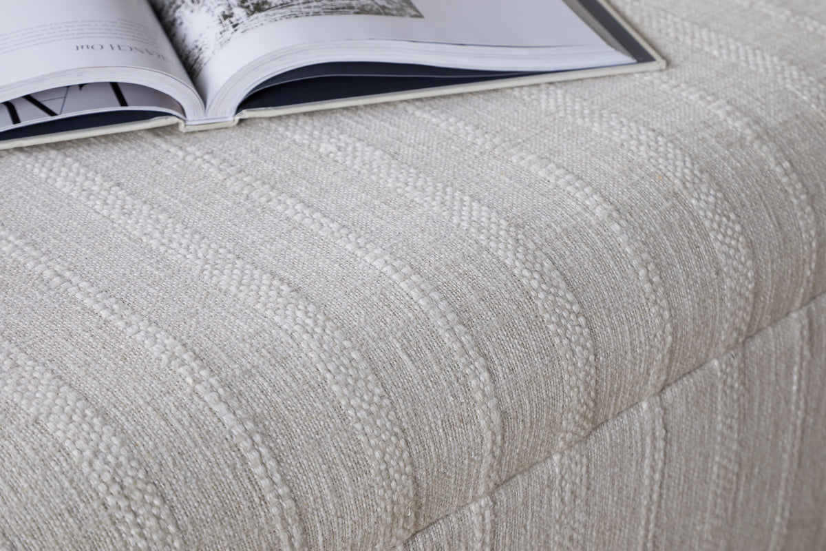 A close-up of the nkuku Kiana Upholstered Storage Ottoman in Natural, featuring a cream-striped, textured fabric and cushioned top, with an open magazine resting on it—an inviting accent for any living space.