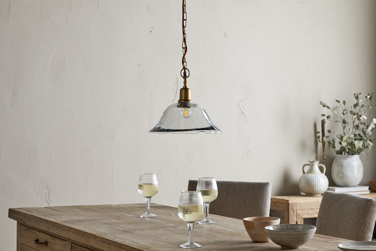 A rustic wooden table holds three glasses of white wine, a small bowl, and a wooden dish. Above, the nkuku Keju Organic Shape Recycled Glass Pendant in clear hangs from a chain. Neutral pottery and greenery decorate the sideboard against a beige wall.