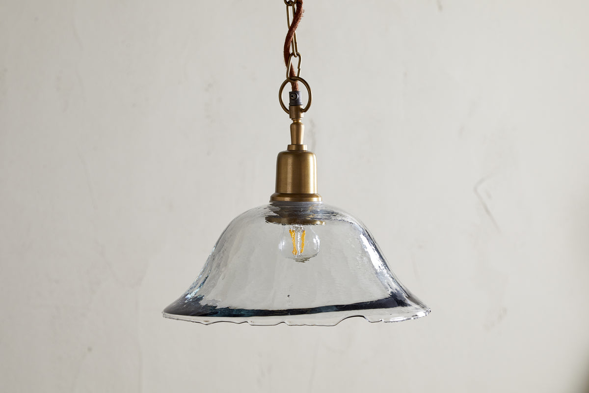 The Keju Organic Shape Recycled Glass Pendant in clear by nkuku features a stylish glass shade suspended from a chain, bringing elegant flair to any room.