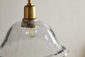A close-up of the Keju Organic Shape Recycled Glass Pendant - Clear by nkuku shows its wavy, transparent glass shade and antique brass top, with a small LED filament bulb inside, emphasizing the lamp’s unique texture and eco-friendly materials.