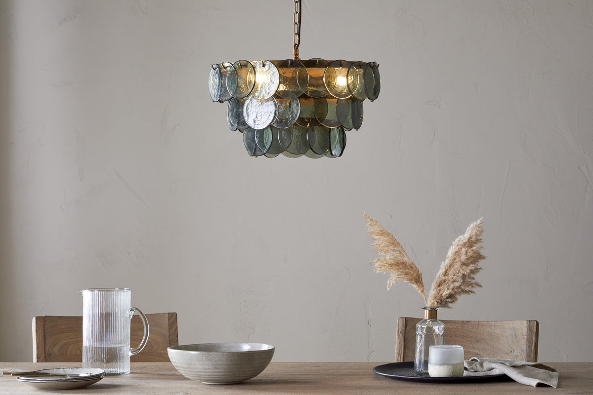 The nkuku Kagitha Recycled Glass Chandelier - Green, featuring layered glass and subtle brass touches, hangs above a minimalist wooden dining table, with its translucent green discs adding elegance to the light-colored wall.