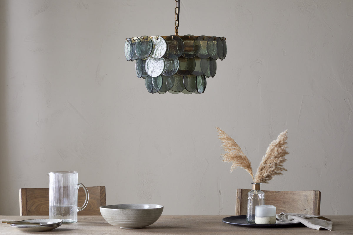 A rustic dining table features neutral ceramic dishes, a glass pitcher, and dried pampas grass in a vase. Overhead, nkuku’s Kagitha Recycled Glass Chandelier in Green hangs, its layered glass and brass detailing set against a textured beige wall.