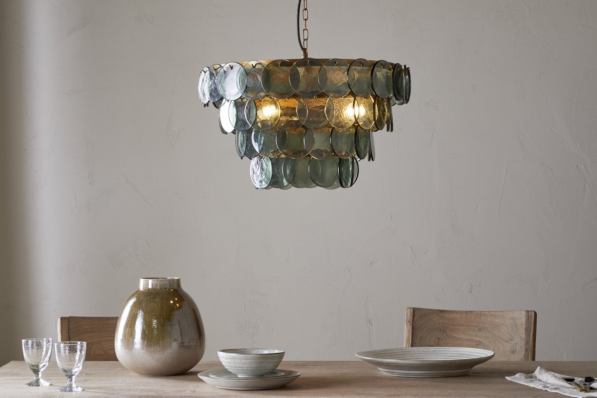 A modern dining table with two wooden chairs, set with a beige vase, stacked ceramic bowls and plates, two clear glasses, and a cloth napkin. Above hangs the nkuku Kagitha Recycled Glass Chandelier in green, softly illuminating the scene.