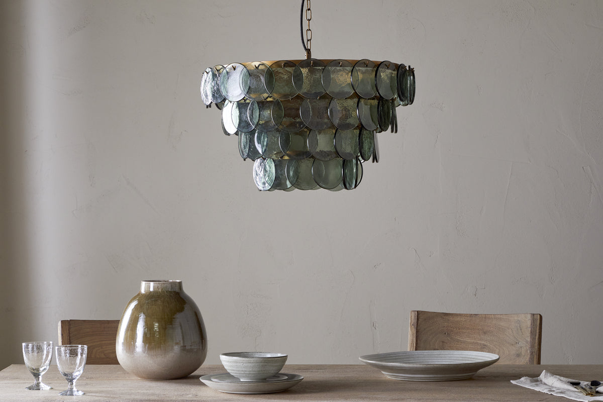 A rustic dining table with light wood chairs holds ceramic dishes, two clear glasses, and a large beige vase. Above hangs the nkuku Kagitha Recycled Glass Chandelier - Green, its layered design casting a soft, elegant glow.