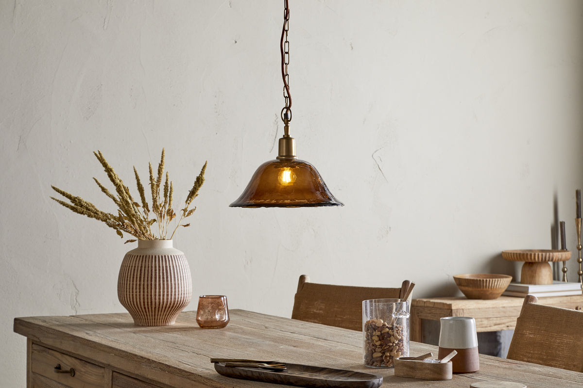 A rustic dining table sits beneath the nkuku Keju Organic Shape Recycled Glass Pendant in Dark Amber, its warm glow highlighting a ribbed vase of dried grasses, small bowls, glassware, and a wooden tray in a softly lit, minimalist room.