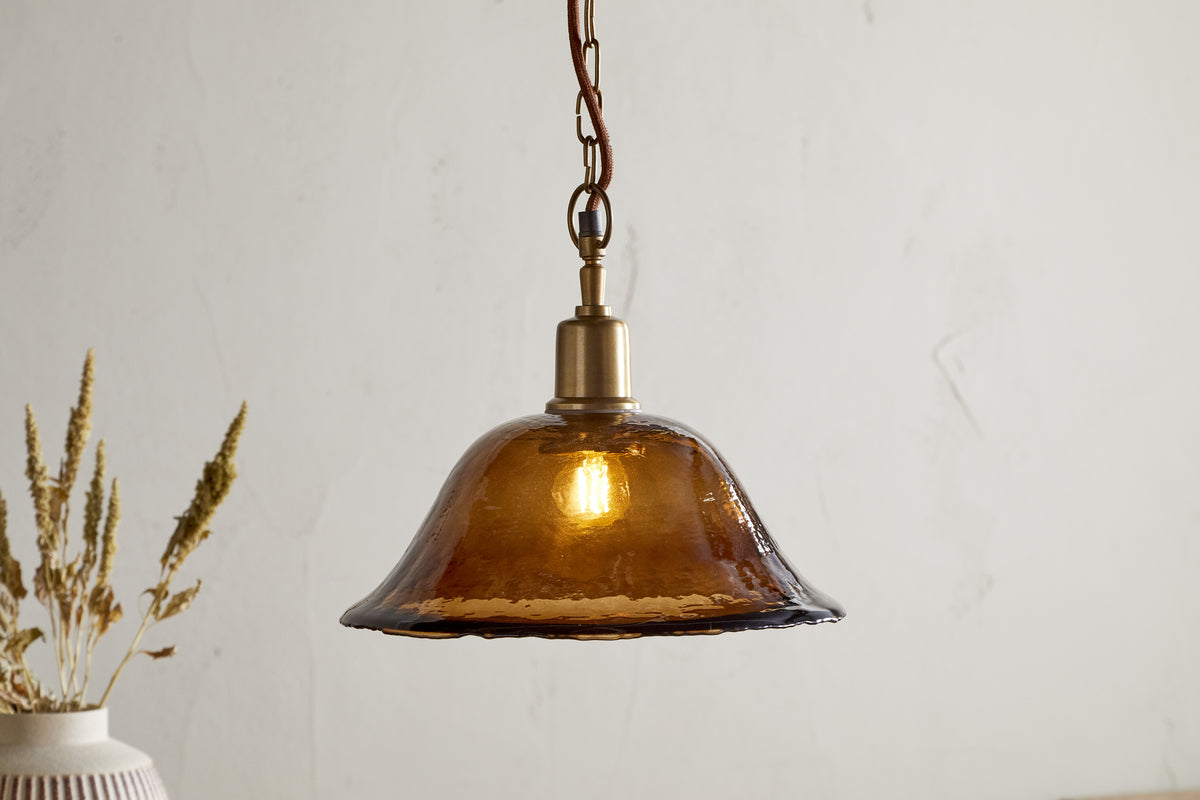The Keju Organic Shape Recycled Glass Pendant in dark amber by nkuku features a visible bulb and antique brass fixture suspended from a chain. In the background, dried flowers in a vase rest against a light-colored wall.