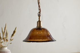 The Keju Organic Shape Recycled Glass Pendant in Dark Amber by nkuku hangs from a chain above a table with a vase of dried wheat, set against a plain, light-colored wall.