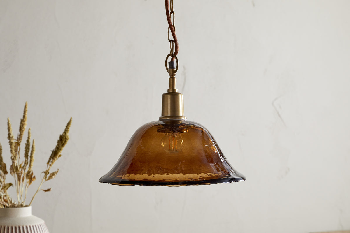 The Keju Organic Shape Recycled Glass Pendant in Dark Amber by nkuku hangs from a chain above a table with a vase of dried wheat, set against a plain, light-colored wall.