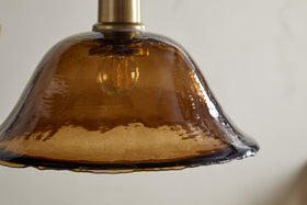 Close-up of the nkuku Keju Organic Shape Recycled Glass Pendant in Dark Amber, showcasing its textured, translucent shade and antique brass fixture against a plain light background.