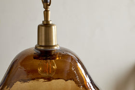Close-up of the nkuku Keju Organic Shape Recycled Glass Pendant in dark amber, featuring an antique brass fixture and a wavy, translucent recycled glass shade that reveals the bulb inside against a neutral background.