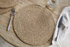 A Keso Woven Placemat by nkuku sits on a light wooden table, paired with minimalist tableware and a glass of water on a handwoven coaster in the upper right corner.