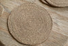 The nkuku Keso Woven Placemats - Brown (Set of 4) rest on a slatted wooden surface, with another placemat and handwoven coasters in view, adding minimalist style to your tableware.