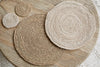 Elevate your minimalist tableware with the Keso Woven Placemats - Brown (Set of 4) by nkuku—handwoven round placemats in varying sizes, beautifully arranged on a light wooden slatted table.